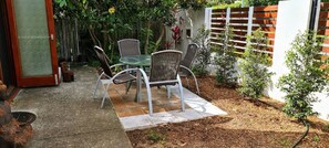 Un patio al aire libre con una mesa de vidrio y sillas de mimbre, rodeado de un jardín con una valla de madera y plantas pequeñas.