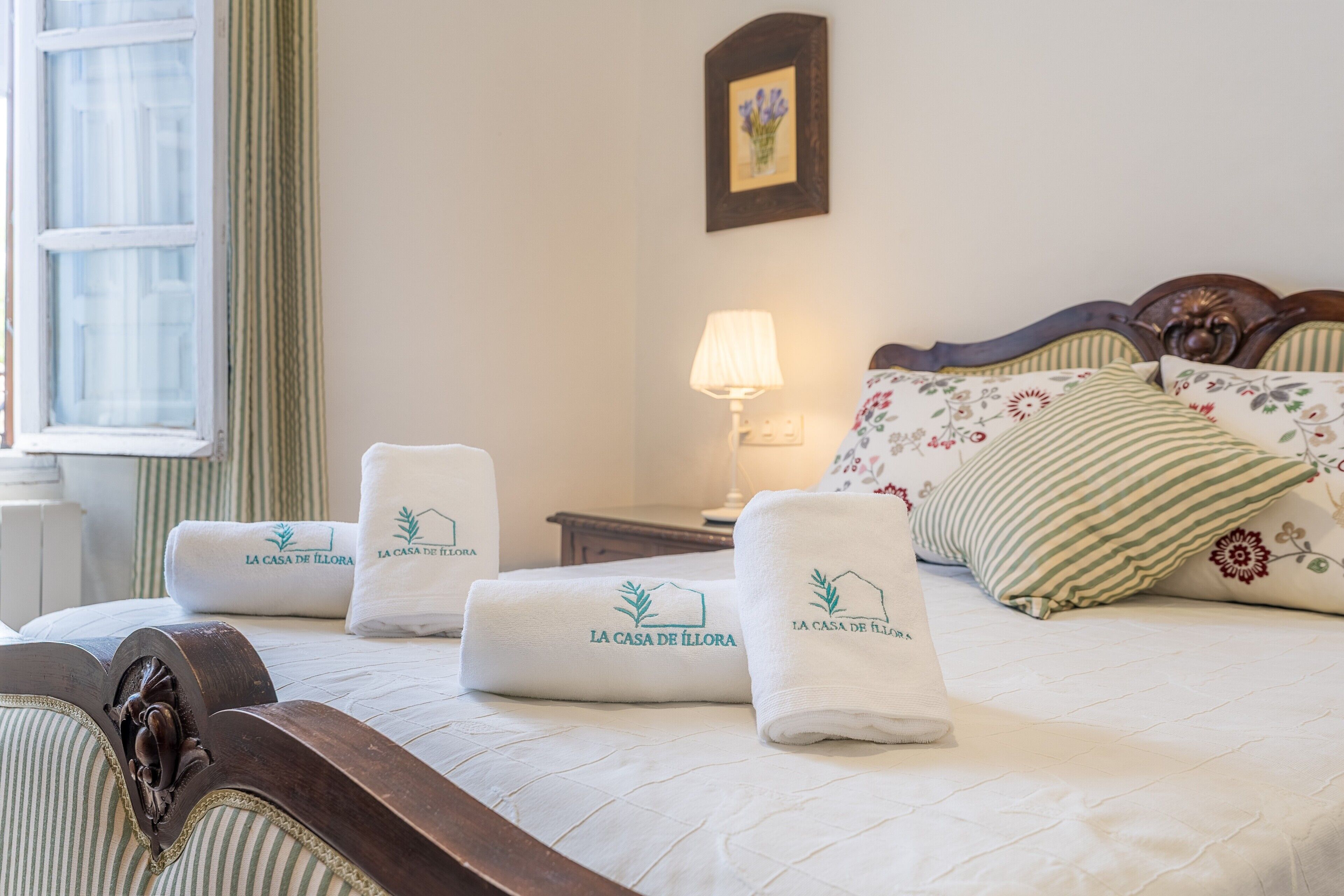 A neatly made bed with floral pillows and a headboard, a bedside lamp, and two rolled towels with the hotel logo.