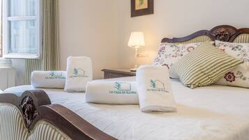 A neatly made bed with floral pillows and a headboard, a bedside lamp, and two rolled towels with the hotel logo.