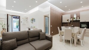 A modern living room with a sofa, dining table, chairs, and a kitchen area with cabinets and a stove.