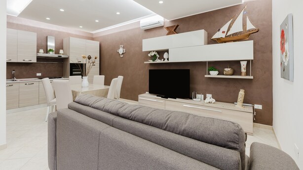 A modern living room with a flat-screen TV, grey sofas, a kitchen area, and a mounted sailboat decoration.