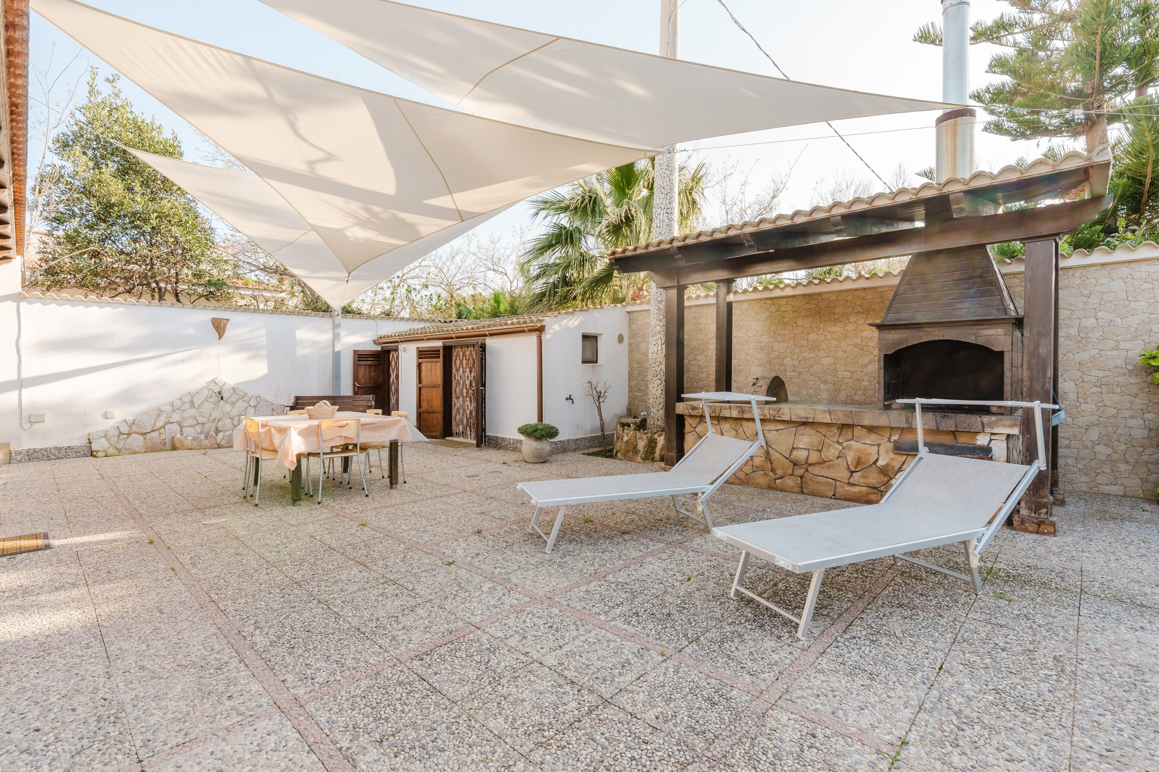 An outdoor patio with a table and chairs, two lounge chairs, a stone oven, and a pergola providing shade.