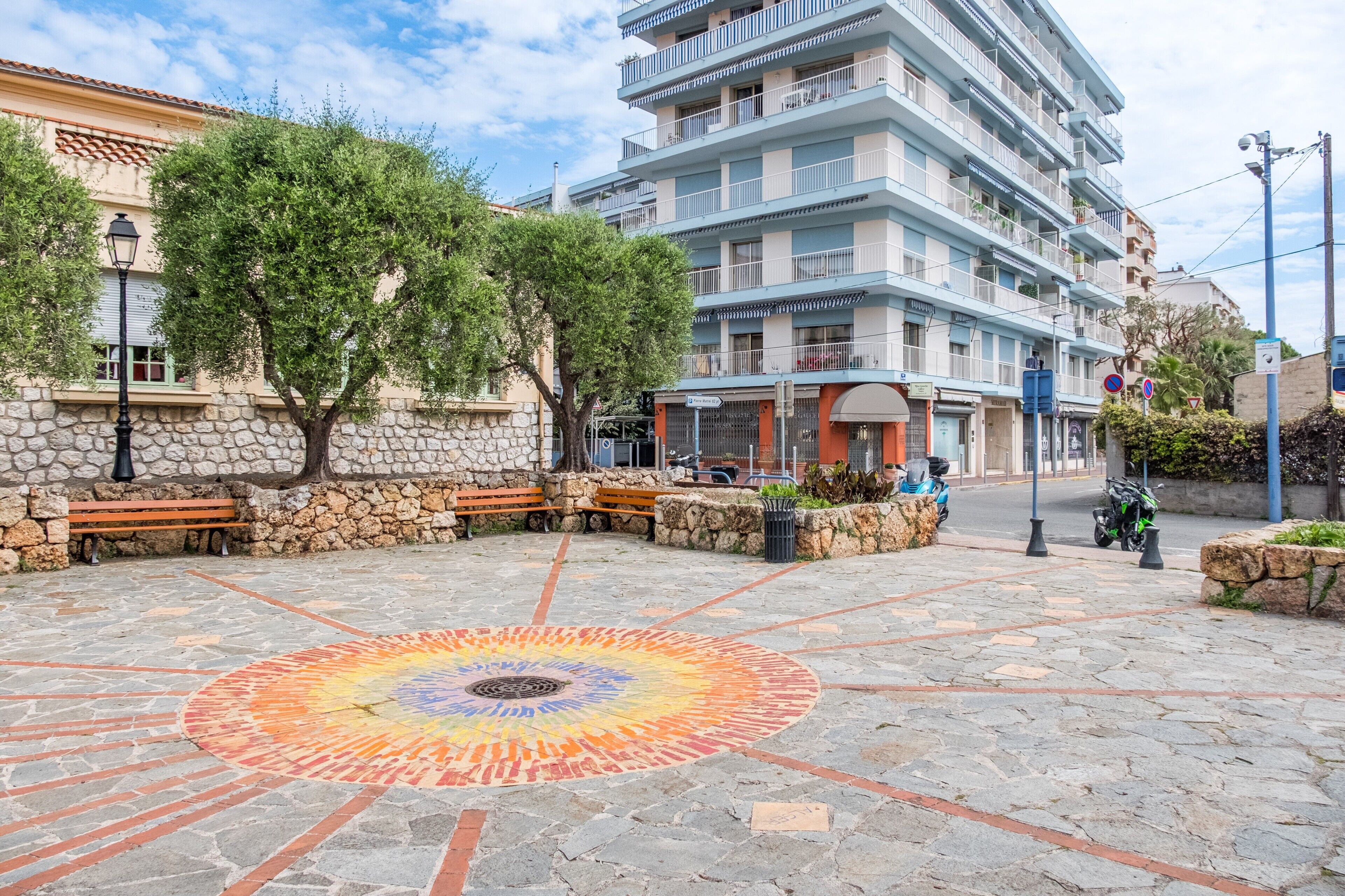 A stone-paved plaza with a colorful circular design, stone benches, and a multi-story building with balconies.