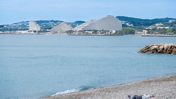 Vista costera con edificios modernos, una playa de guijarros y agua tranquila.