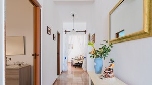 Un couloir avec une porte en bois, une console blanche surmontée d’un vase et d’une statuette, un miroir, et un lit visible au loin.