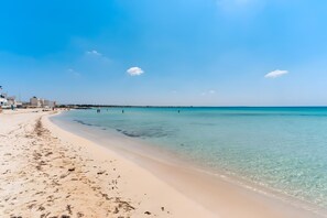 Ein Strand mit klarem Wasser, einem sandigen Ufer und Gebäuden im Hintergrund.