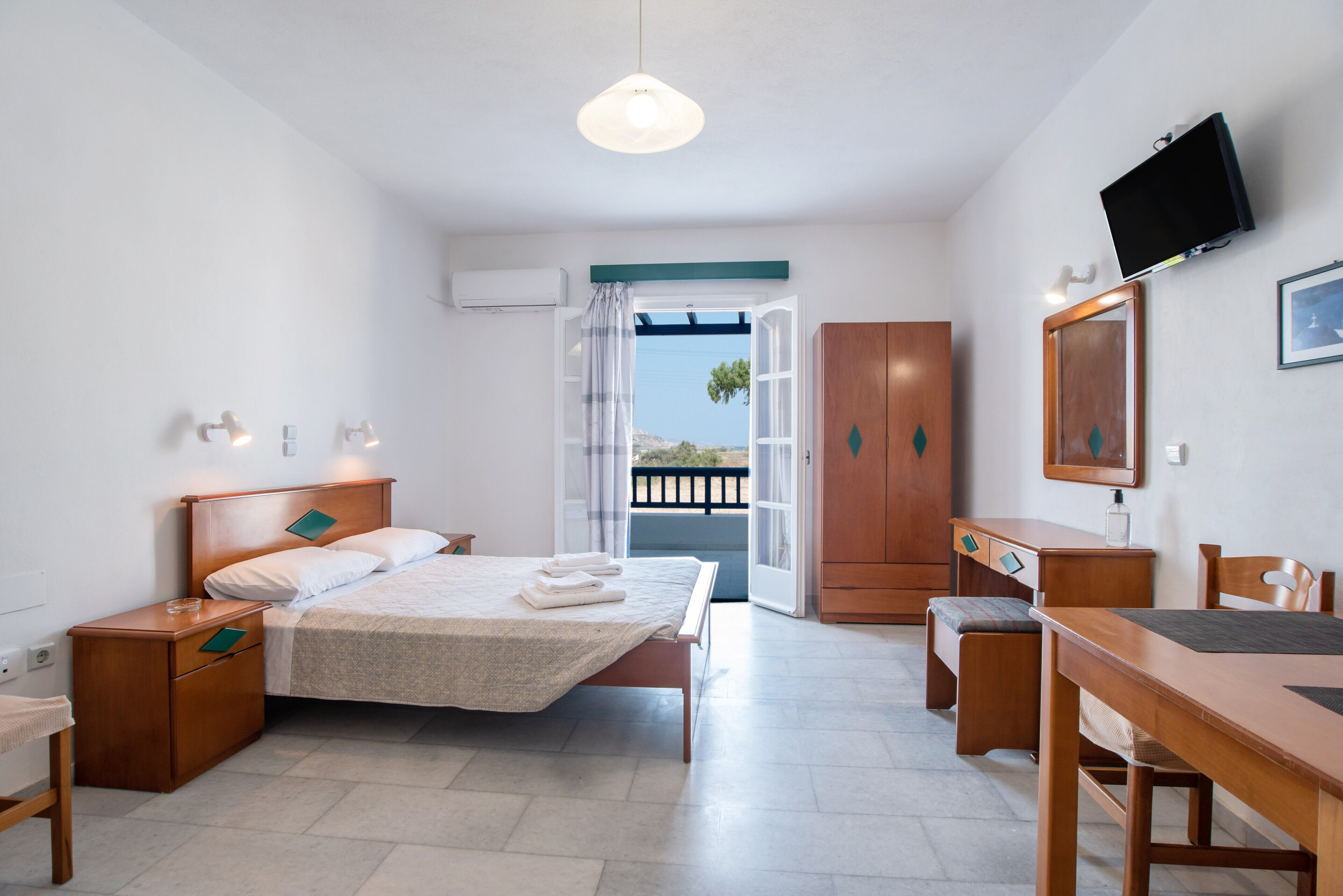 A bedroom with a bed, bedside tables, a desk, a chair, a television, and a view through an open door.