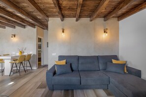 A modern living room with a sectional sofa, wooden ceiling beams, and a dining area with chairs and a table.
