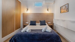 A bedroom with a wooden headboard, a bed with dark blue bedding, two bedside tables, and a small window with blinds. - Holiday Home 'Miranda 2' with Sea View, Wi-Fi and Air Conditioning (Igueste de Candelaria)