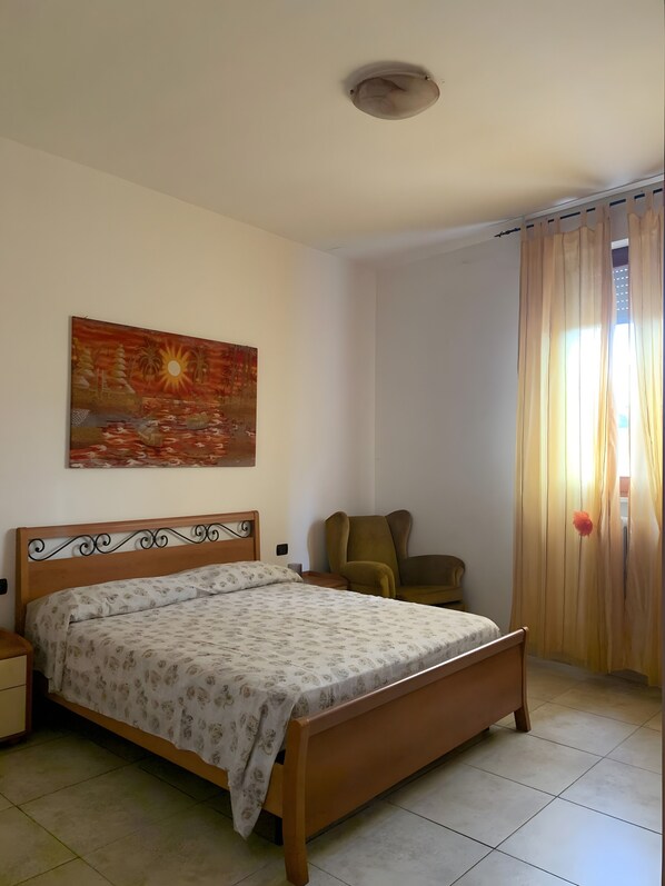 A bedroom with a bed, a chair, a painting, and a window with curtains.