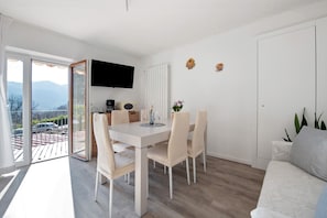 A modern dining area with a white table and chairs, a flat-screen TV mounted on the wall, and a view of the outdoors through a sliding glass door.