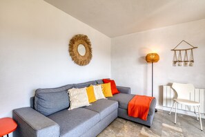Un divano angolare grigio con una coperta rossa, una poltrona bianca, una lampada da terra e un elemento decorativo a parete.