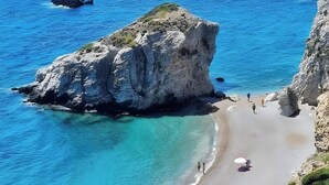 A coastal beach with clear turquoise waters, rocky cliffs, and a few umbrellas on the sand.