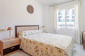 Un dormitorio con cama, mesitas de noche, un adorno en la pared, una ventana con cortinas y vistas a edificios.
