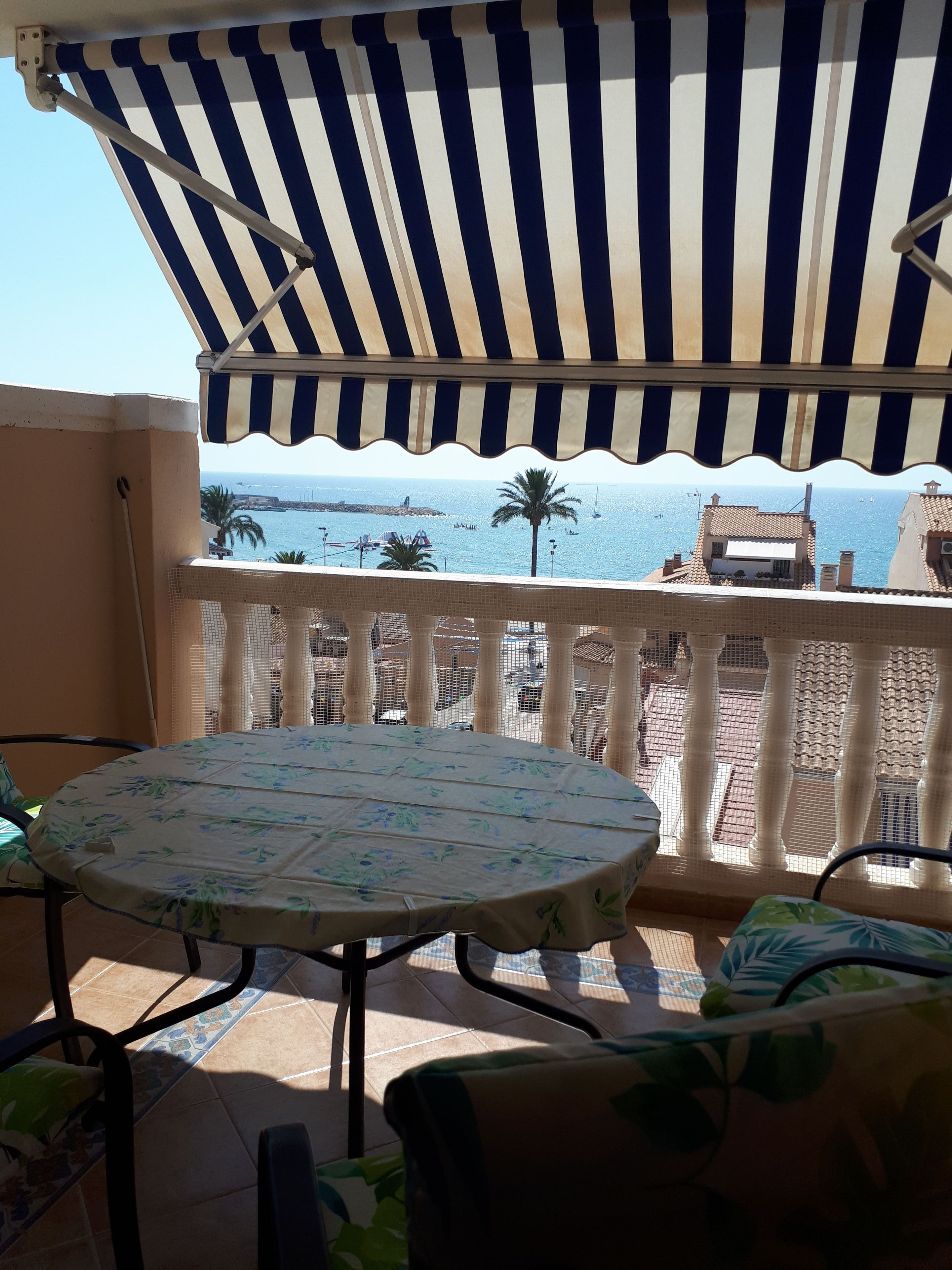 A balcony with a striped awning, a round table with a floral tablecloth, and a view of the sea and buildings.