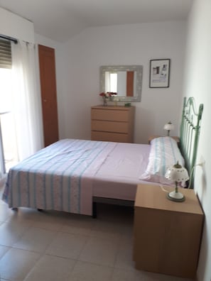 Ein Schlafzimmer mit einem Bett, einem Schminktisch, einem Nachttisch, einer Lampe, einem Spiegel und einem Fenster mit Vorhängen.
