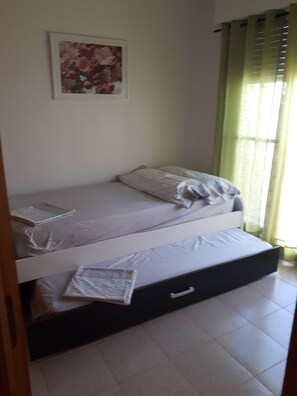 A single bed with a mattress, pillows, and a blanket. There is a small bedside table with a lamp. A framed picture hangs on the wall above the bed.