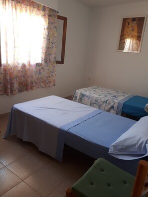 Ein Zimmer mit zwei Betten, einem grünen Sessel und einem Fenster mit blumigen Vorhängen.