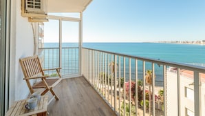 A balcony with a wooden chair, a table with a mug, and a view of the sea and buildings.