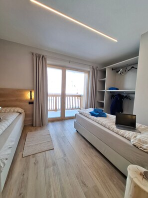 A modern hotel room with a bed, a sofa bed, a laptop, a wardrobe, and a balcony with curtains.