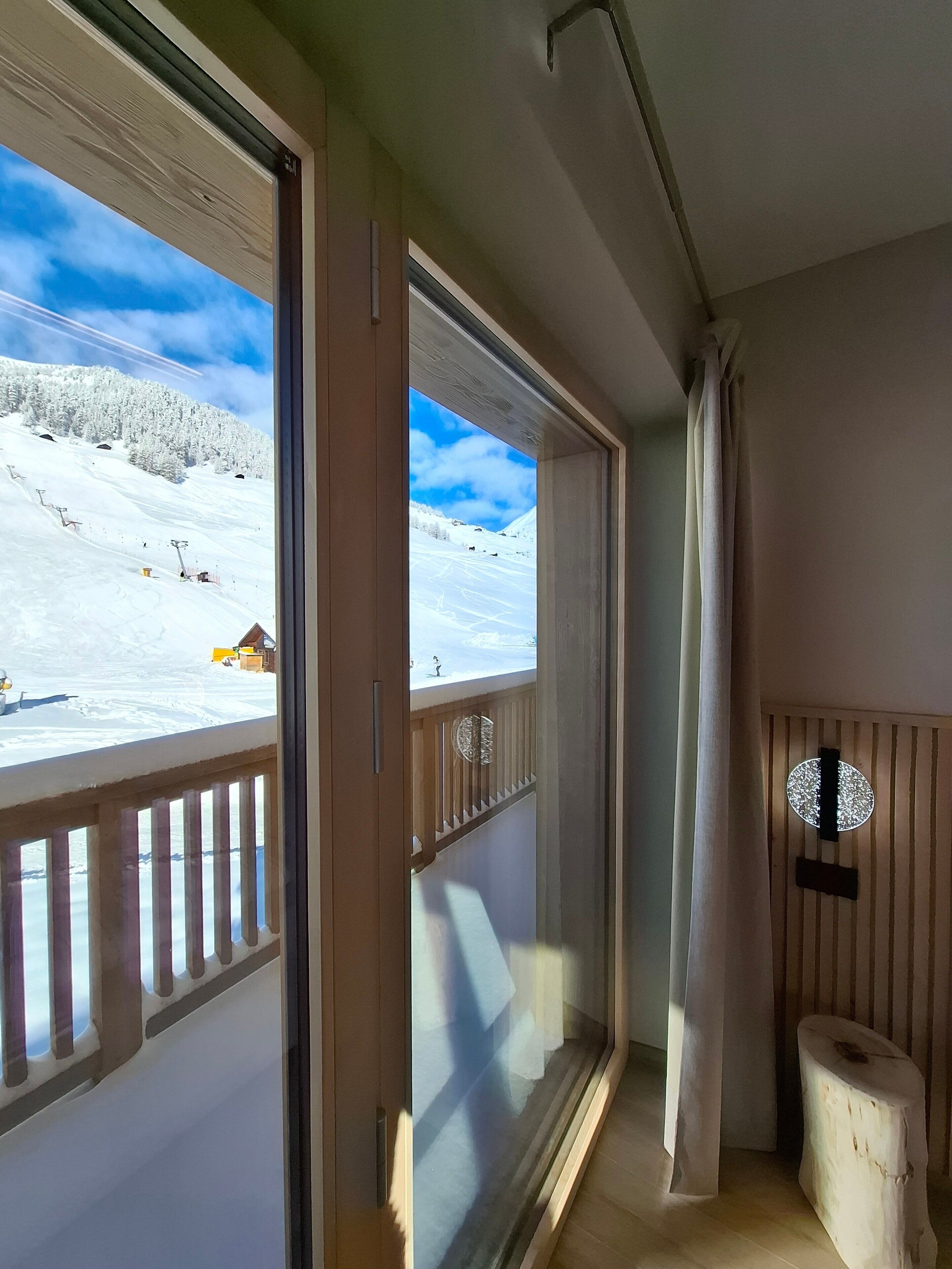 A room with a large window overlooking a snowy mountain landscape, a wooden railing, and a small wooden stool.