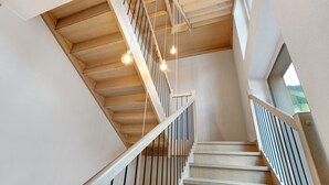 Una escalera de madera con barandilla, techo con vigas a la vista y lámparas colgantes.