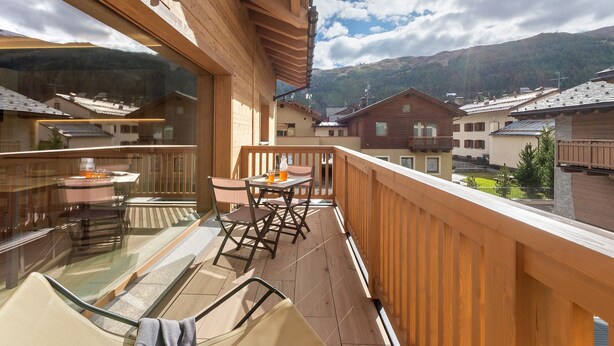 Un balcón de madera con una mesa y sillas de vidrio, con vista a un pueblo montañoso.