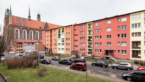 A multi-story residential building with a red and yellow facade, a parking lot with several cars, and a historic church with a red roof in the background.
