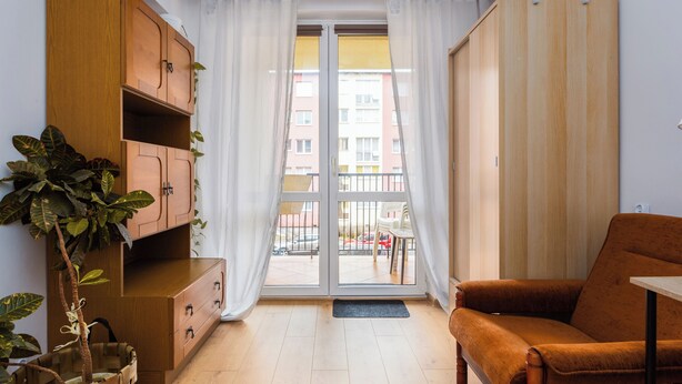 A room with a wooden cabinet, a brown sofa, a potted plant, and a sliding glass door leading to an outdoor balcony.
