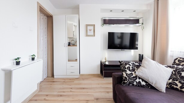 A modern living room with a wooden floor, a dark sofa with patterned cushions, a flat-screen TV mounted above a dark wooden console, and a white wardrobe with a mirror. - Park Grabiszyński Standard Studio (Wrocław)
