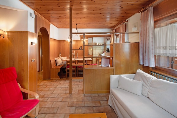 A compact living space with a wooden ceiling, a sofa, a red chair, a kitchen area, and a dining table.