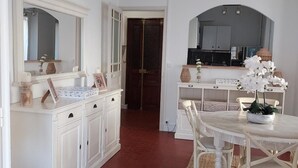 Une pièce avec une coiffeuse blanche, une table de salle à manger ronde entourée de chaises, un luminaire suspendu et un coin cuisine visible à travers une porte ouverte.