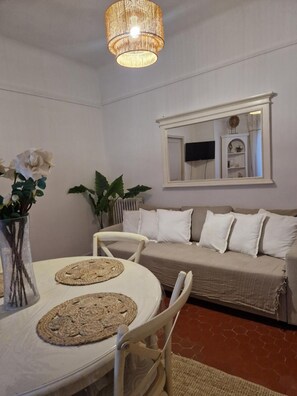 Un coin repas avec une table ronde, deux sets de table en fibres naturelles, des chaises blanches, un canapé beige, un grand miroir et un luminaire suspendu.
