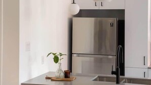 Cozinha moderna com geladeira de aço inoxidável, armários brancos e um balcão com duas cadeiras azuis.