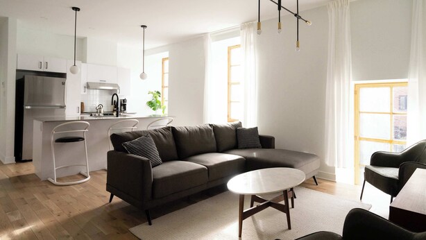 A modern living room with a grey sofa, a round coffee table, and a kitchen area with a fridge and sink.