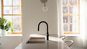 A modern kitchen with a white countertop, a black faucet, and a pendant light. There are two windows with wooden frames and a potted plant on the left.
