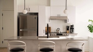 A modern kitchen with a stainless steel refrigerator, a gas stove, and a coffee maker. There are three white chairs with black cushions.