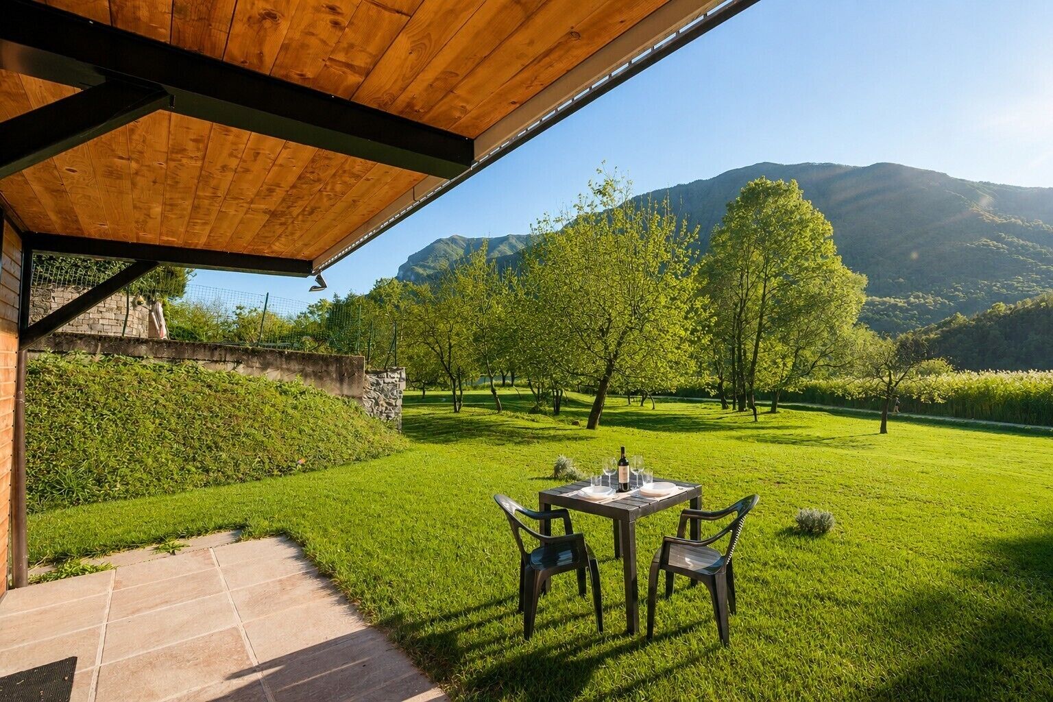 A covered patio with a table set for two, overlooking a well-maintained lawn and distant mountains.