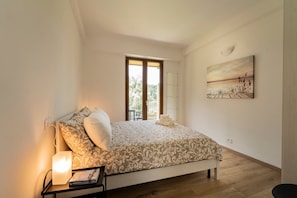A bedroom with a bed, a bedside table with a lamp, a painting on the wall, and a window with a view of greenery.