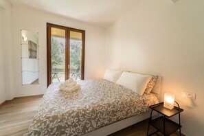 A bedroom with a bed, a bedside table with a lamp, a mirror, and a view of the outdoors.