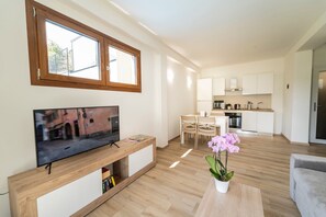 A modern living room with a flat-screen TV, wooden furniture, a dining area, and a kitchen with white cabinets.