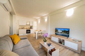 Uma sala de estar moderna com um sofá, mesa de centro e televisão. Cozinha visível ao fundo, com área de refeições.