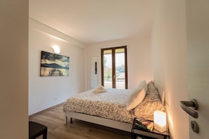 A bedroom with a bed, a nightstand, a lamp, a painting, and a view through a door. - Holiday Apartment "7" with Lake View, Shared Pool & Wi-Fi (Carlazzo)