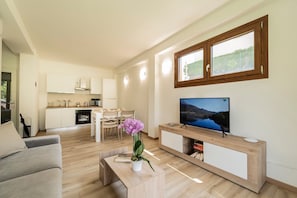 A modern living room with a flat-screen TV, a wooden coffee table with a plant, a sofa, and a dining area with a kitchen in the background.