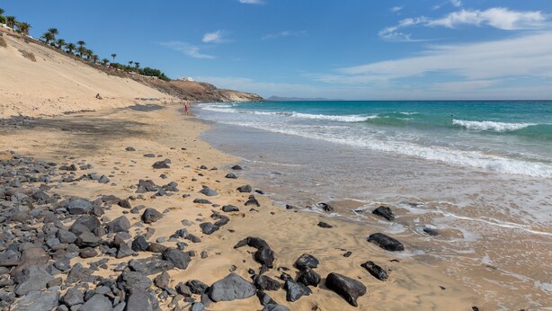 A beach with rocky terrain, a clear sky, and a distant building on the left.