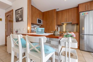A modern kitchen with wooden cabinets, a stainless steel refrigerator, a microwave, and a dining area with white chairs and a blue table.