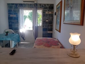 Una habitación con pared de piedra, una mesa con mantel azul, una cama con colcha estampada, una lámpara de pie y un cuadro en la pared.