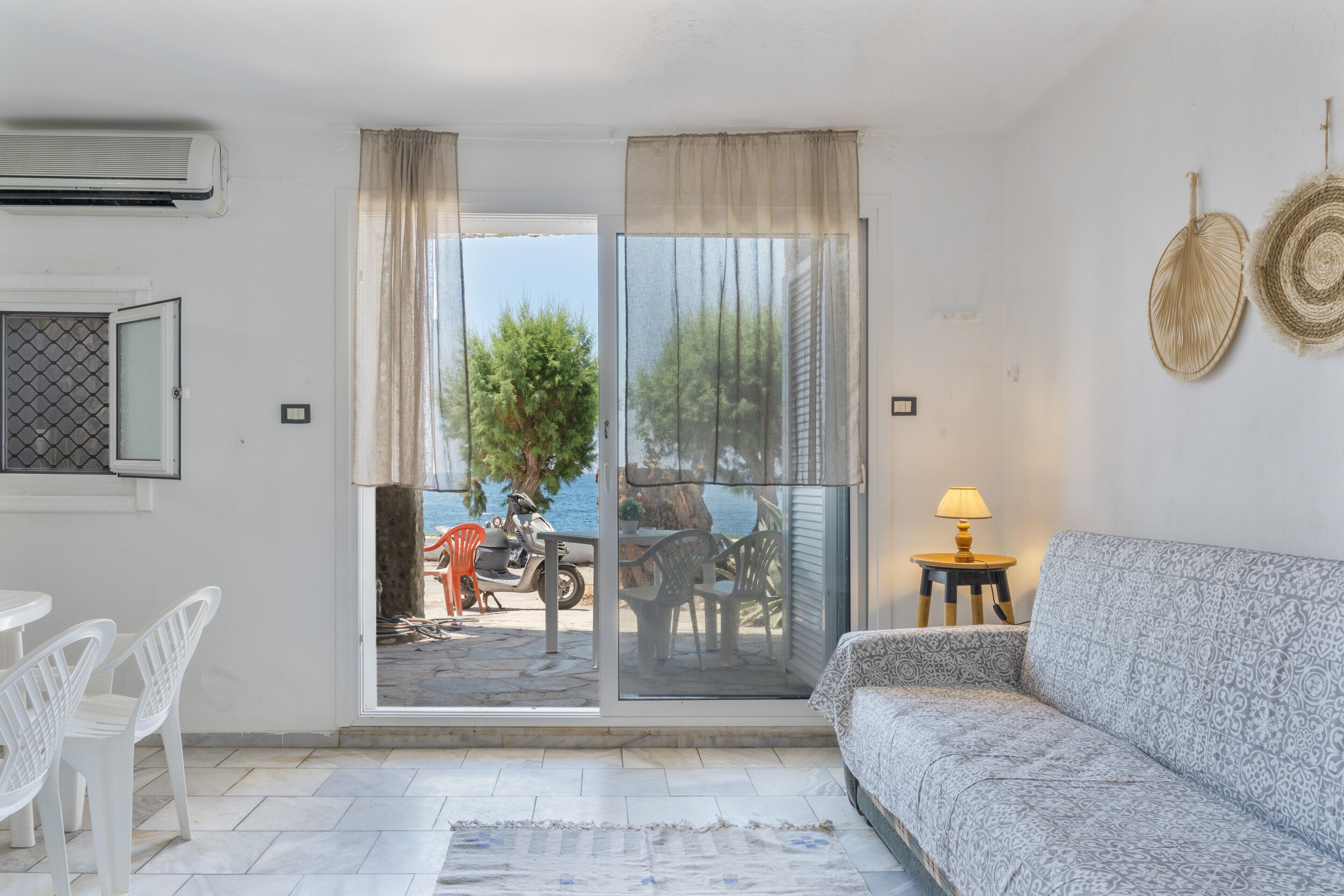 Une pièce comprenant un canapé, une table avec des chaises, une lampe et une vue sur l’extérieur, accessible par une porte vitrée, donnant sur un espace extérieur où se trouvent une moto et un arbre.