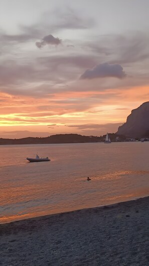 A sunset over a calm sea with a boat and a sailboat, a beach, and a cliff in the background.
