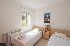 Habitación de hotel con una cama individual, una mesita de noche pequeña con lámpara, un radiador y una ventana con vistas al verdor.